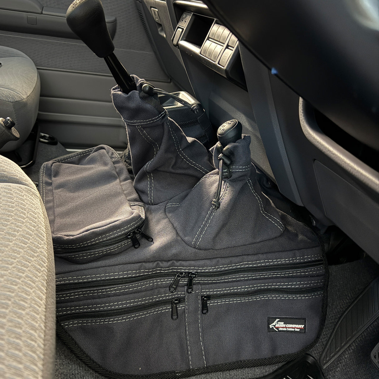 A charcoal car interior accessory with multiple zippered pockets and looped handles, placed on the footwell inside a vehicle. A small brand patch is visible on the lower right.