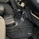 A charcoal car interior accessory with multiple zippered pockets and looped handles, placed on the footwell inside a vehicle. A small brand patch is visible on the lower right.