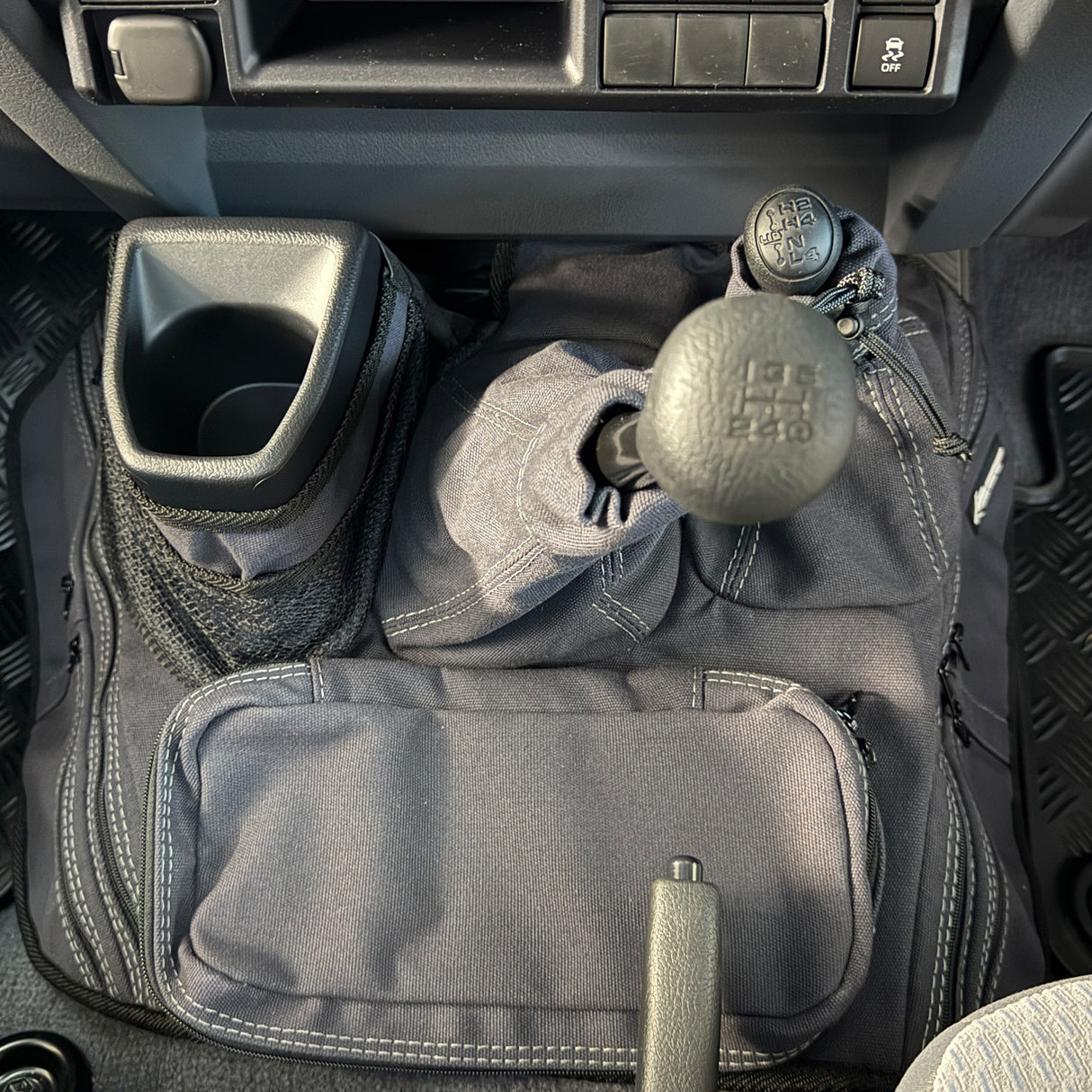 A charcoal car interior footwell organizer mat with multiple pockets, mesh storage, and a cup holder, shown installed around a manual gear shift and handbrake.