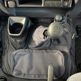 A charcoal car interior footwell organizer mat with multiple pockets, mesh storage, and a cup holder, shown installed around a manual gear shift and handbrake.