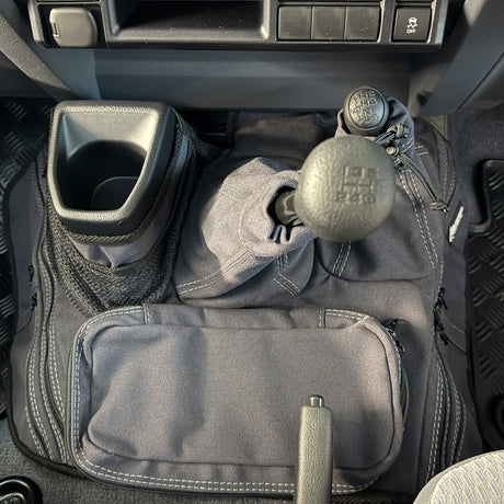 A charcoal car interior footwell organizer mat with multiple pockets, mesh storage, and a cup holder, shown installed around a manual gear shift and handbrake.