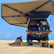 Person setting up a campsite on a beach with a vehicle having an extended awning.