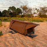 A brown camping swag pitched on sandy ground outdoors, with shrubs, trees, and a cloudy sky in the background.