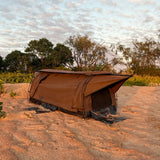 A brown camping swag pitched on sandy ground outdoors, with trees and shrubs in the background under a cloudy sky.