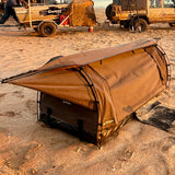 A brown canvas camping swag with a black base and black frame, set up on sandy ground with off-road vehicles in the background.