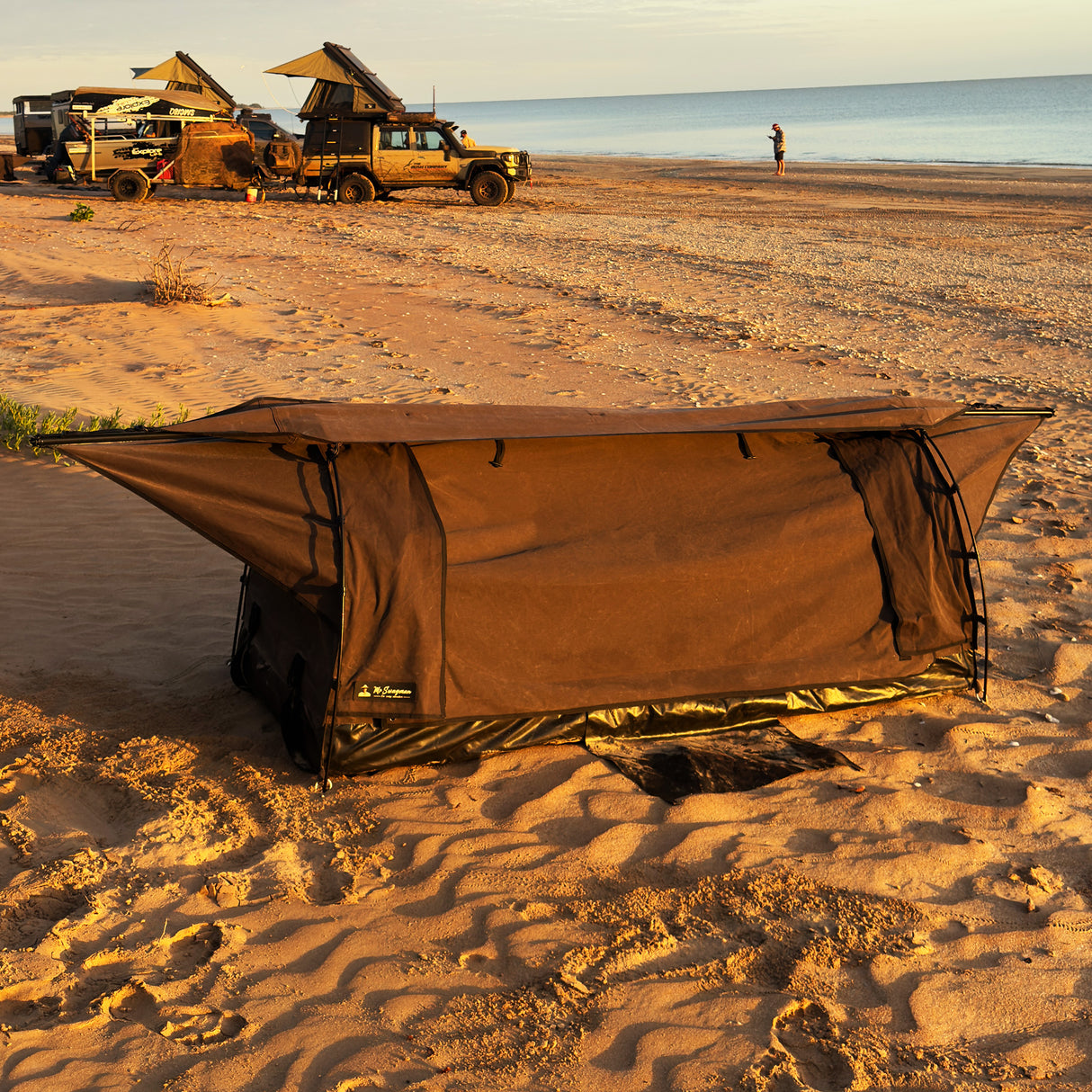 A brown camping swag is pitched on a sandy beach at sunset, with desert vehicles and the ocean visible in the background.