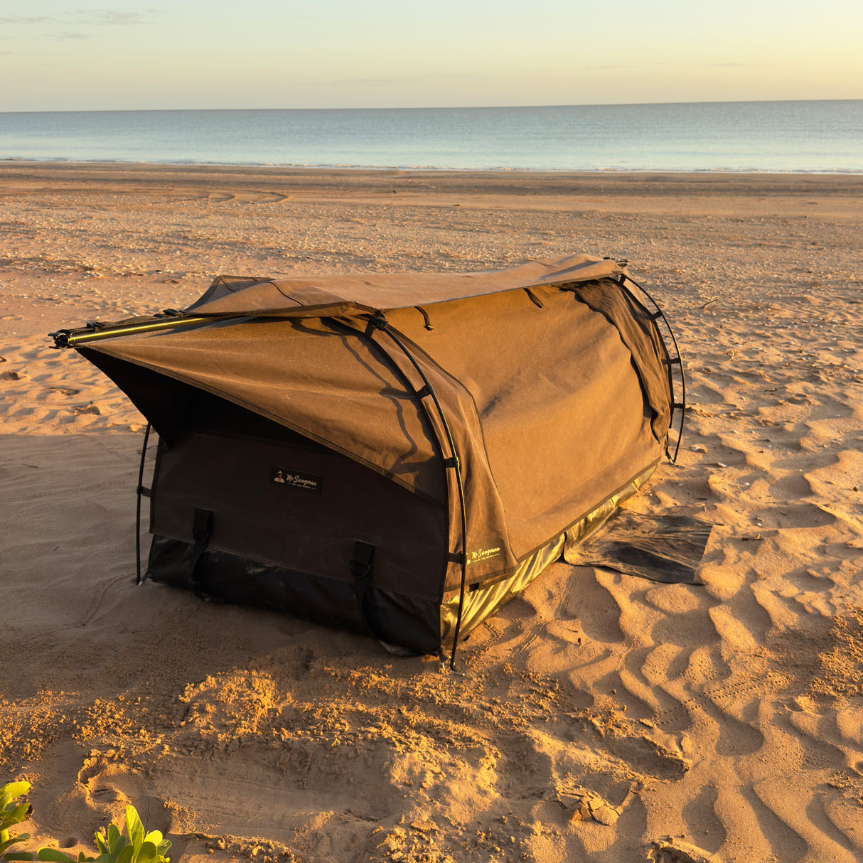 A brown swag set up on sandy beach ground with the ocean and sky in the background.