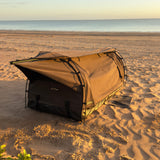 A brown swag set up on sandy beach ground with the ocean and sky in the background.