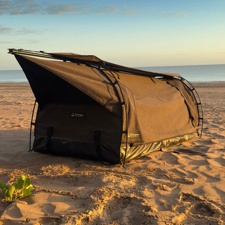 A brown camping swag with an attached awning is pitched on a sandy beach with the ocean and sky in the background.