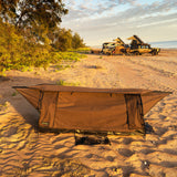 A brown camping  swag shelter set up on sandy beach ground, with trees, vehicles, and ocean visible in the background at sunset.