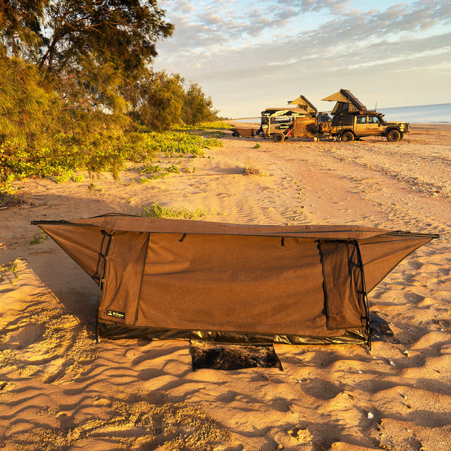 A brown camping  swag shelter set up on sandy beach ground, with trees, vehicles, and ocean visible in the background at sunset.