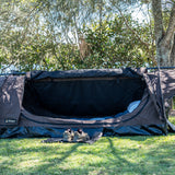A brown camping swag set up on a grassy lawn outdoors, with hiking boots placed in front of the open entrance and trees in the background.