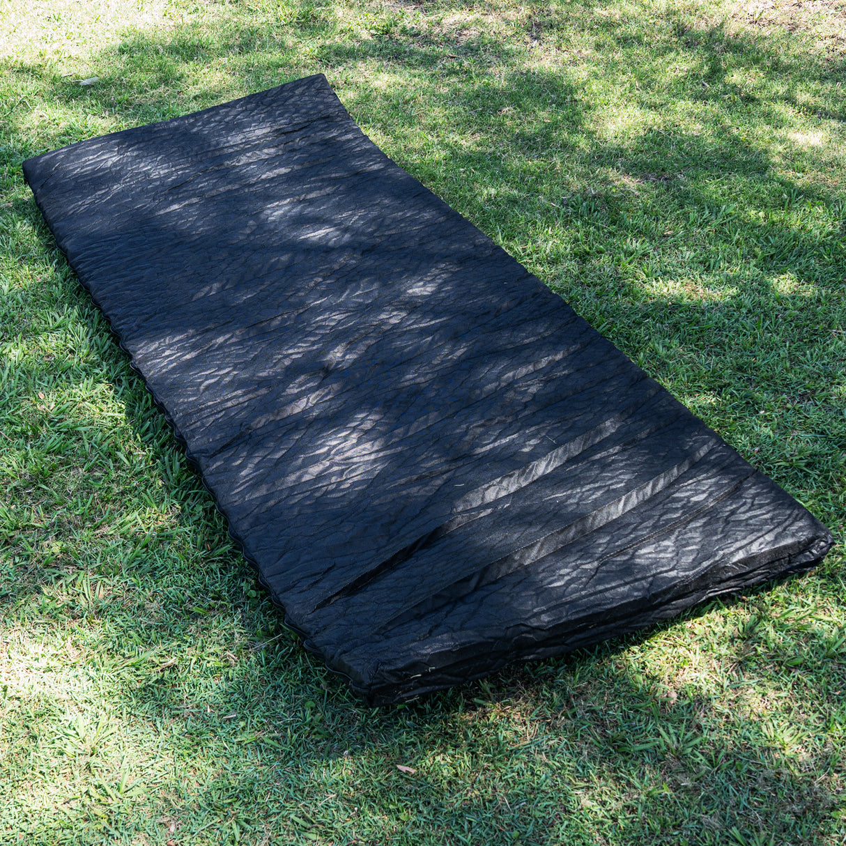 A black camping mat or sleeping pad laid out on green grass outdoors, with dappled sunlight and shadows across the surface.