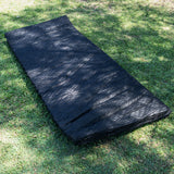 A black camping mat or sleeping pad laid out on green grass outdoors, with dappled sunlight and shadows across the surface.