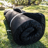 A rolled-up black swag mattress secured with white straps, lying on green grass outdoors with trees and sunlight in the background.