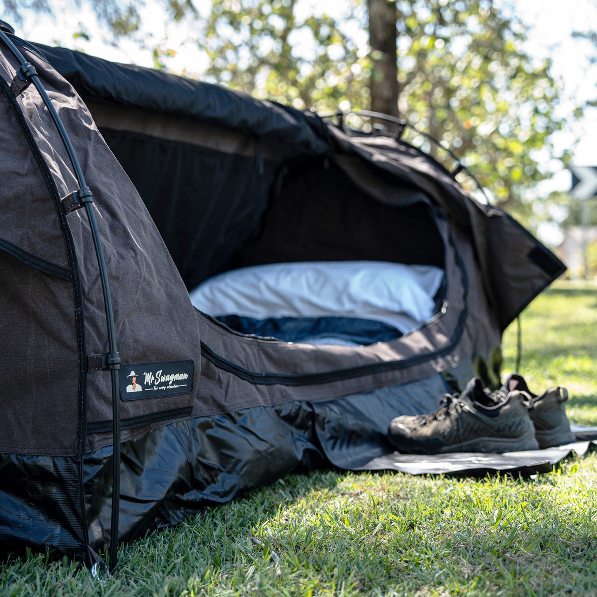 A brown camping swag set up on green grass outdoors, with a white pillow inside and a pair of hiking shoes on a mat nearby.