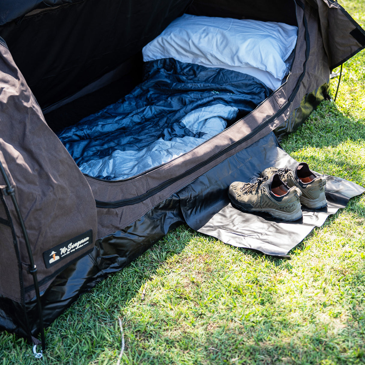 A brown camping swag set up on green grass, with a blue sleeping bag, white pillow, and a pair of hiking shoes placed outside the tent entrance.