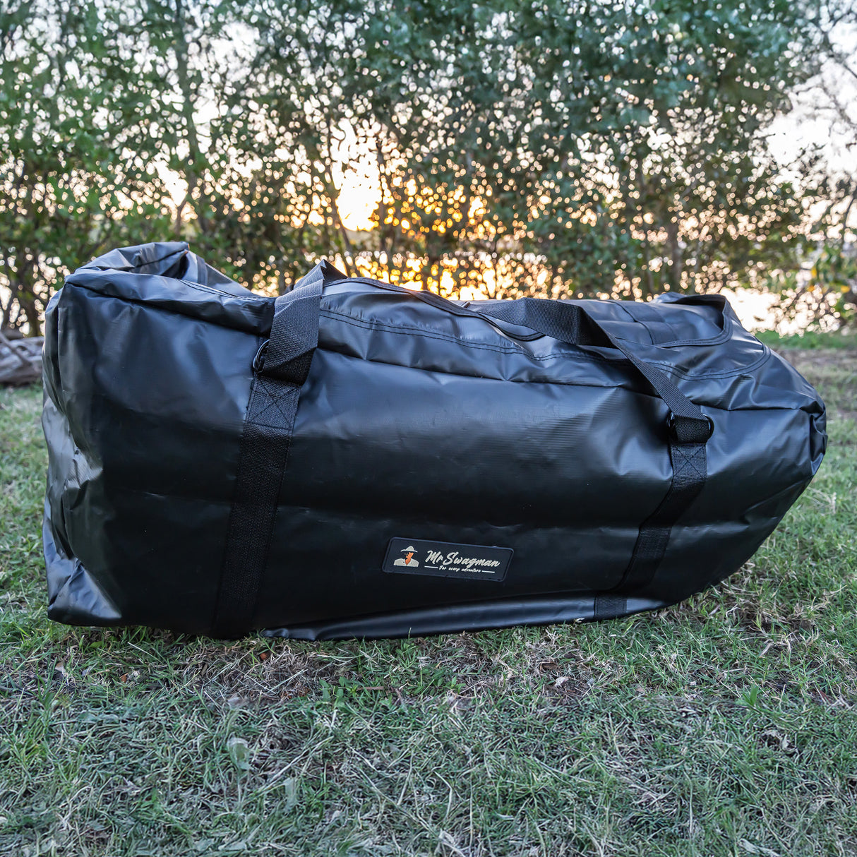 A large black waterproof duffel bag with a small 'The Swagman' brand label, resting on a grassy lawn with trees in the background.