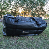 A large black waterproof duffel bag with a small 'The Swagman' brand label, resting on a grassy lawn with trees in the background.
