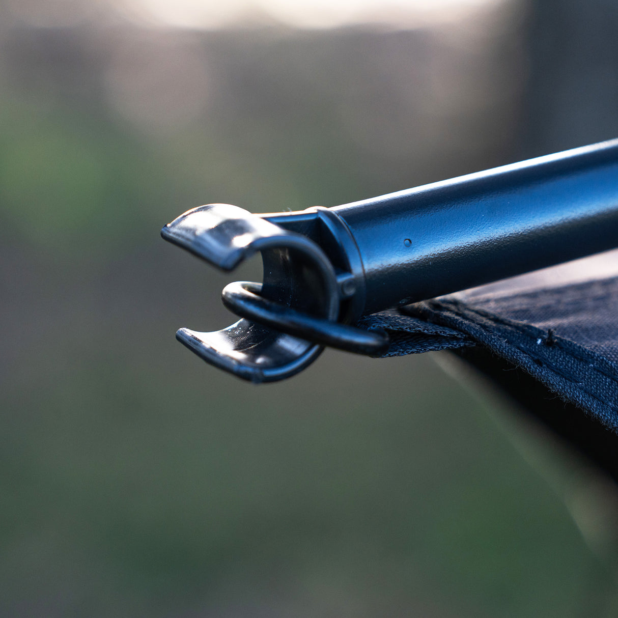Close-up of a black metal tent pole hook inserted into a black fabric canopy, photographed outdoors with a blurred natural background.