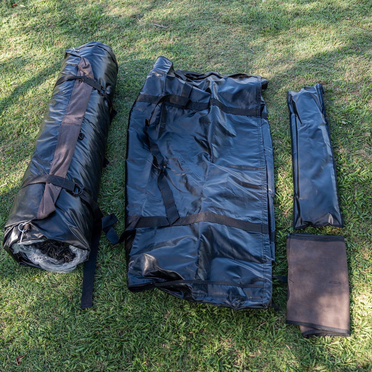 A black camping mat or sleeping pad set laid out on green grass, showing both unpacked and rolled configurations with straps and a brown fabric pad.
