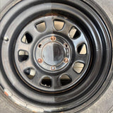 A close-up of a black steel wheel rim and tire, showing the center cap, lug holes, and part of the sidewall.