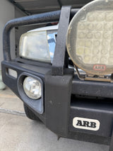 Close-up of a black ARB front bumper and bull bar on an off-road vehicle, showing the main headlight, auxiliary round light, and a white ARB logo badge on the bumper.