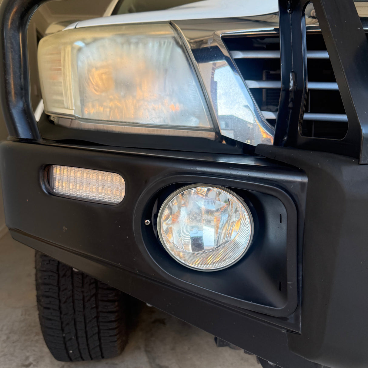 Close-up of the front bumper area of a black off-road vehicle, showing a round auxiliary fog light, a rectangular LED light bar, and part of the tire in a garage setting.