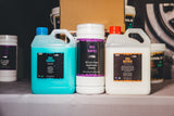 Three automotive care product containers on a shelf: a turquoise jug labeled Big Wash, a white canister labeled Rig Wipes, and a white jug labeled Rig Revive, with additional containers blurred in the background.