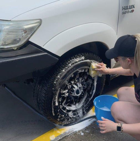 A person crouches beside a white Hilux vehicle, washing the front wheel with a sponge while holding a blue bucket on an outdoor paved surface.