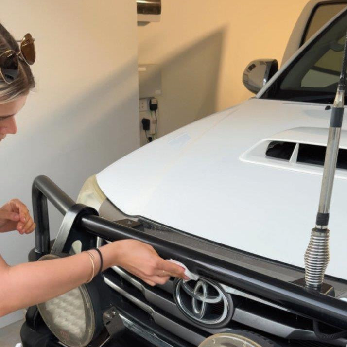 A woman in a garage is shown from the side, touching a black metal front bumper bar mounted across the front of a white Toyota vehicle.