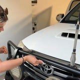 A woman in a garage is shown from the side, touching a black metal front bumper bar mounted across the front of a white Toyota vehicle.